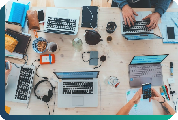 Several laptops on a table. Three people are working. You can also see food, drinks and accessories.