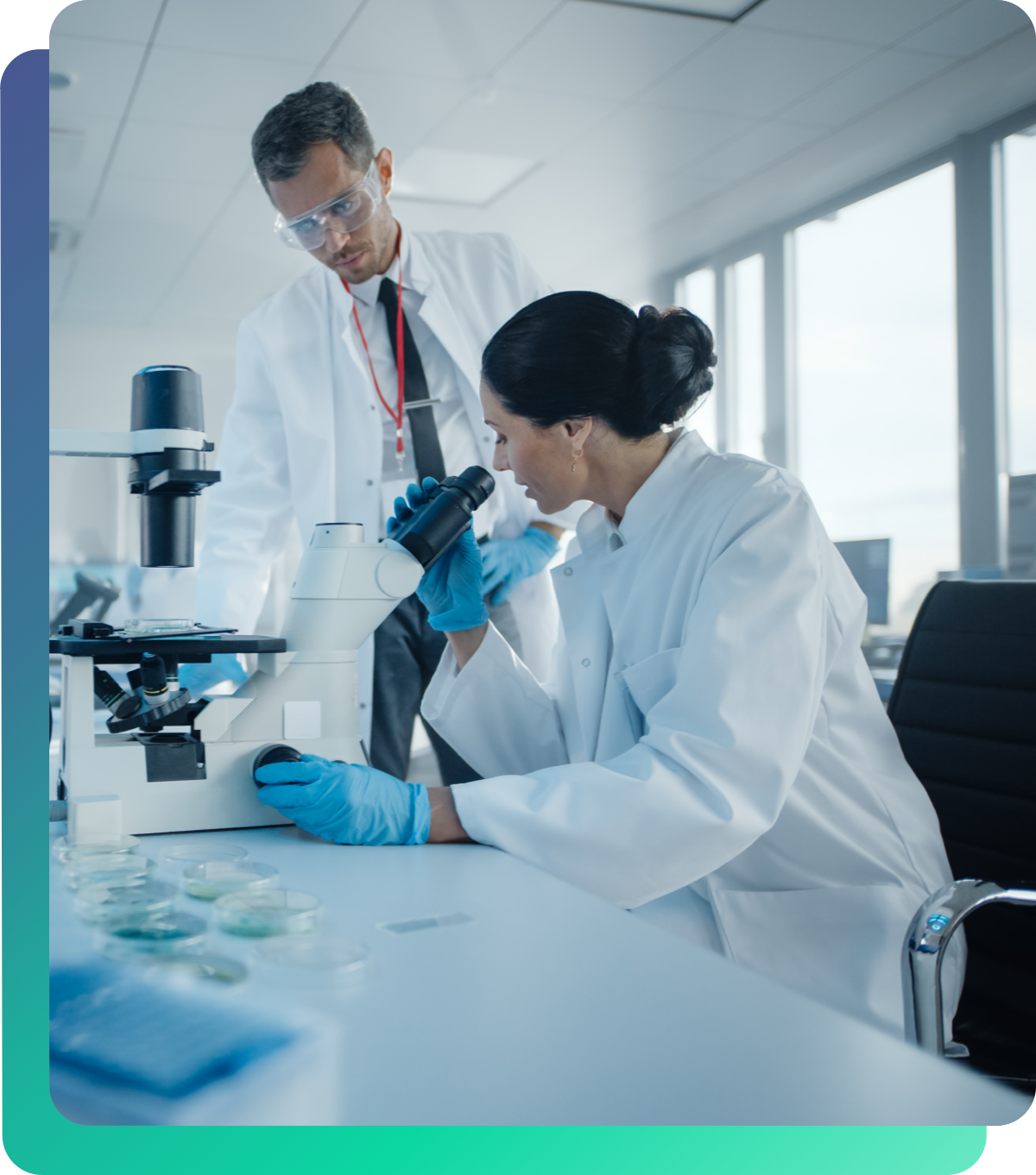 A scientist works at the microscope, a scientist stands next to her and points at something.